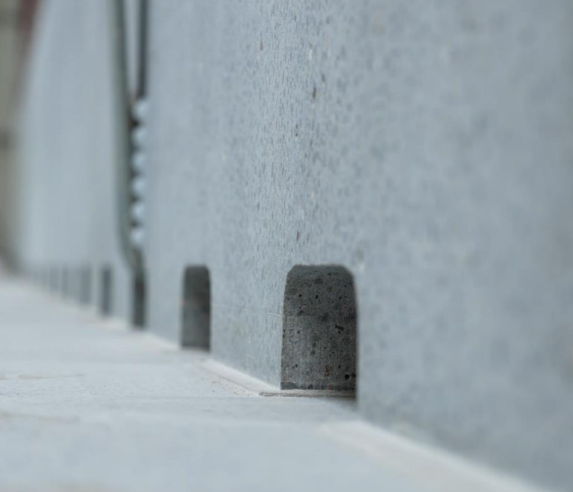 Ein Mitarbeiter in Schutzkleidung und Handschuhen kippt eine Probe frischen Carbonbetons aus einem Eimer in einen zylindrischen Behälter. Der Beton ist zähflüssig und grau, während der Mitarbeiter den Eimer vorsichtig über dem Behälter hält, um den Beton aufzufangen. Die Szene spielt in einer industriellen Umgebung, möglicherweise in einer Produktionshalle oder einem Labor, mit unscharfem Hintergrund, der die Aufmerksamkeit auf die Aktion des Mitarbeiters lenkt.